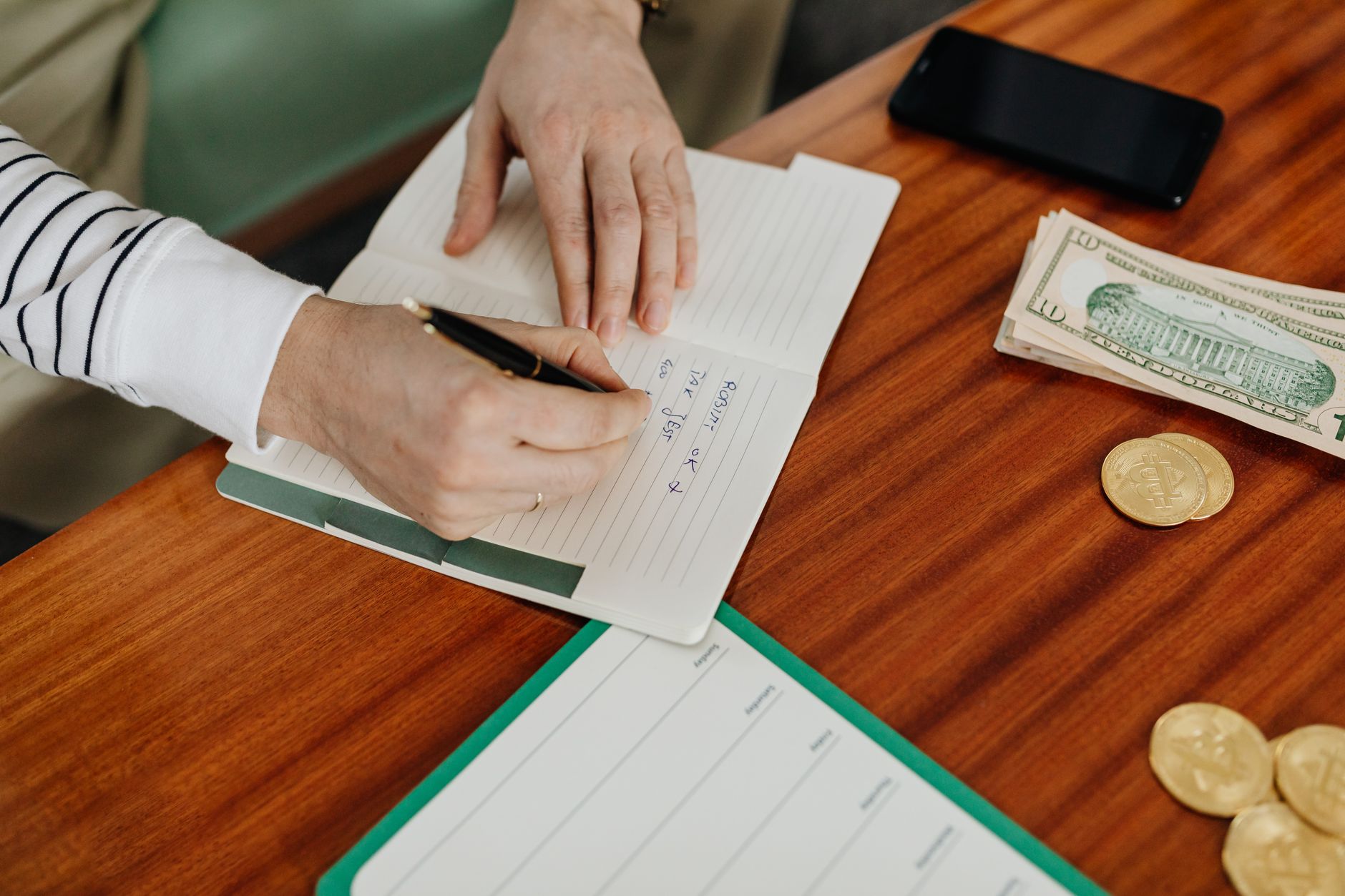 A hand writing in a ledger beside coins